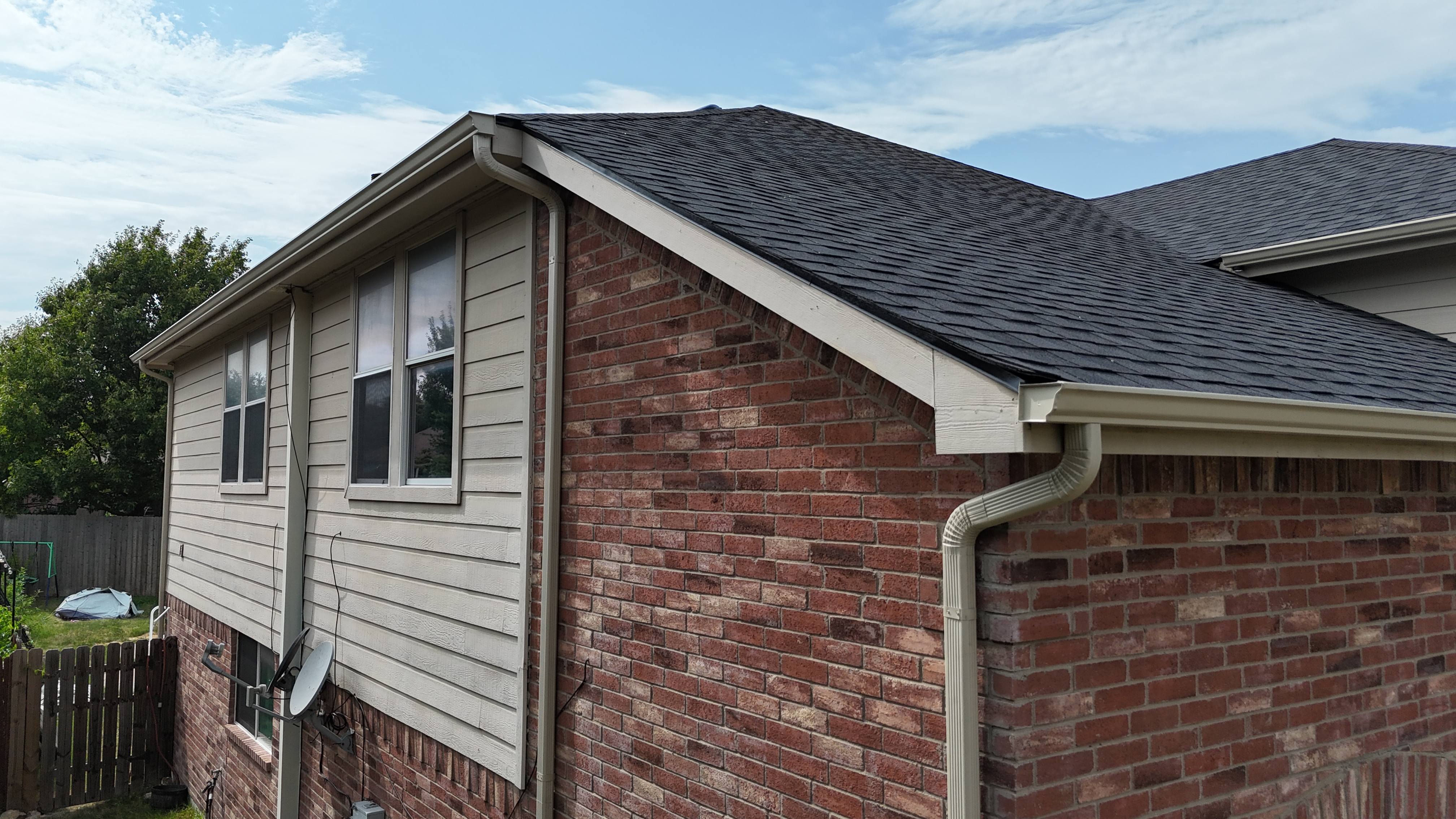 Side view of completed black shingle roof on red brick home with new gutters and downspout